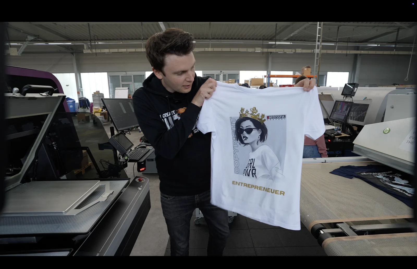 Frank hält in einer Textildruck-Produktionshalle ein weißes T-Shirt mit Design hoch