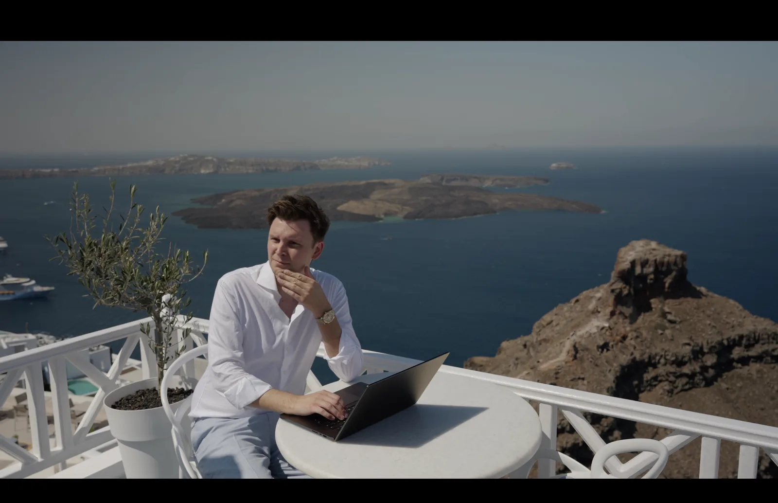 Frank sitzt mit Laptop auf einer weißen Terrasse auf Santorini und blickt aufs Meer